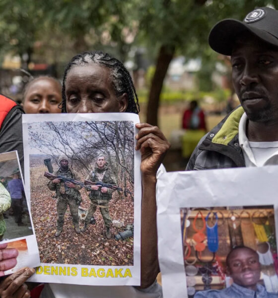 Three adults outdoors hold printed posters showing people’s photos during a daytime vigil or gathering in a park.