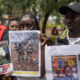 Three adults outdoors hold printed posters showing people’s photos during a daytime vigil or gathering in a park.