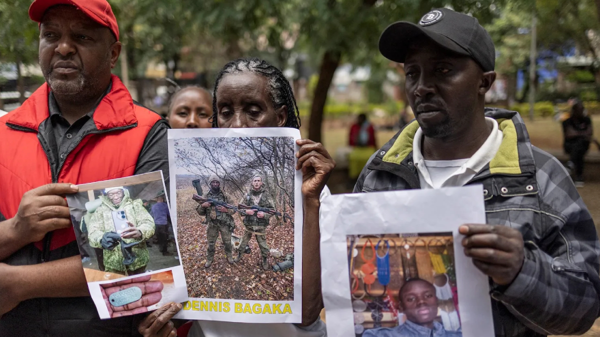 Three adults outdoors hold printed posters showing people’s photos during a daytime vigil or gathering in a park.