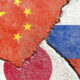 Cracked concrete wall with overlapping flags: China (red with yellow stars) on the left, Japan (red circle on white) at the bottom, and Russia (white-blue-red stripes) on the right, all weathered.