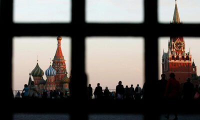 Silhouetted crowd on Red Square at dusk, with Saint Basil's Cathedral and the Spasskaya Tower visible through a window grid.