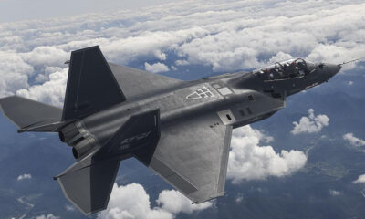 Twin-seat stealth fighter jet in flight above white clouds, seen from behind and slightly below, with dark grey angular wings and twin exhausts visible.",