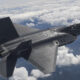Twin-seat stealth fighter jet in flight above white clouds, seen from behind and slightly below, with dark grey angular wings and twin exhausts visible.",