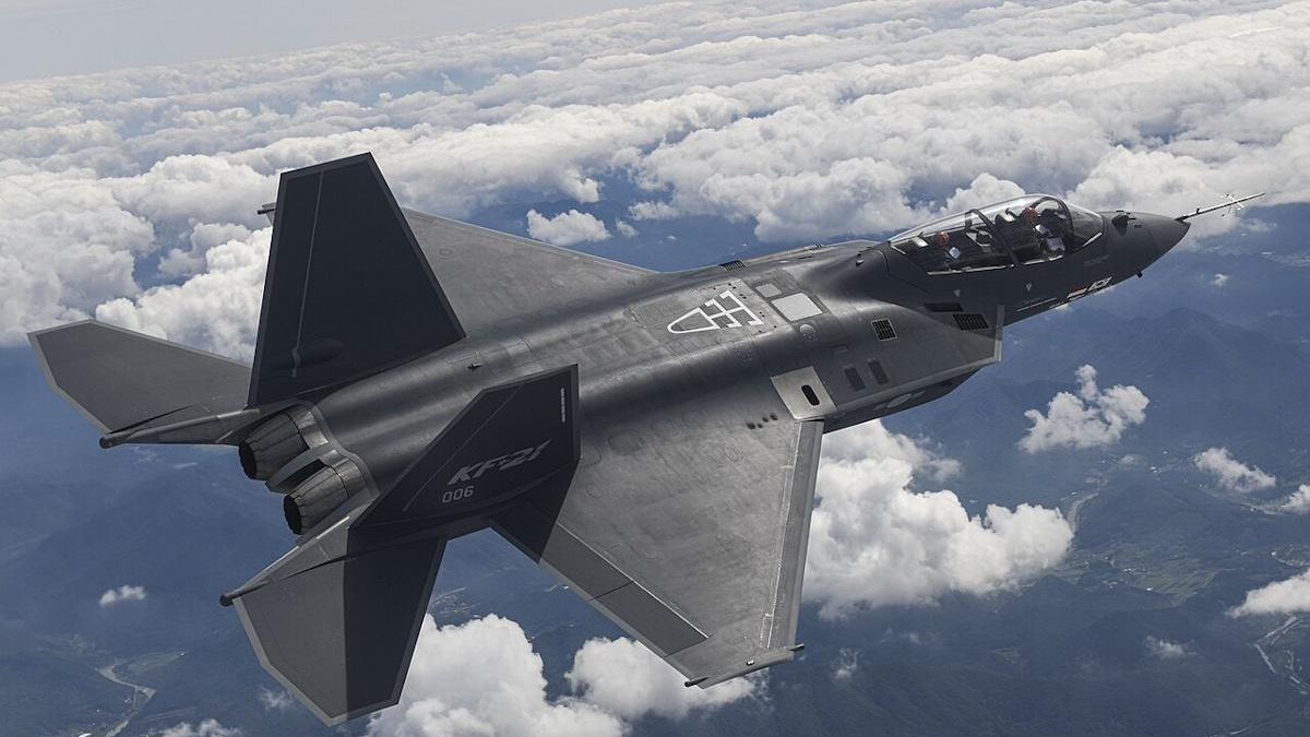 Twin-seat stealth fighter jet in flight above white clouds, seen from behind and slightly below, with dark grey angular wings and twin exhausts visible.",