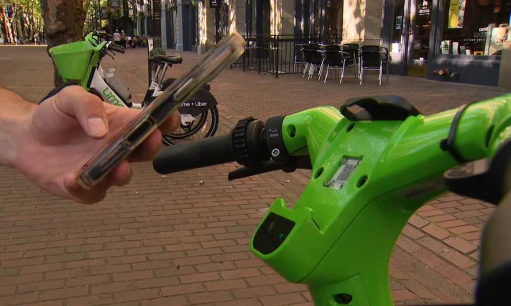 Person holding a smartphone near the handlebar of a bright green Lime scooter on a brick sidewalk with cafe seating in the background, suggesting unlocking the ride.