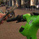 Person holding a smartphone near the handlebar of a bright green Lime scooter on a brick sidewalk with cafe seating in the background, suggesting unlocking the ride.