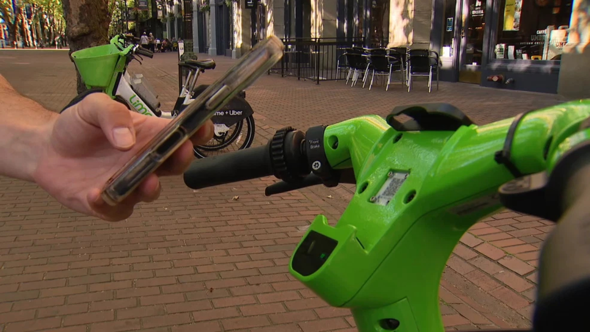 Person holding a smartphone near the handlebar of a bright green Lime scooter on a brick sidewalk with cafe seating in the background, suggesting unlocking the ride.
