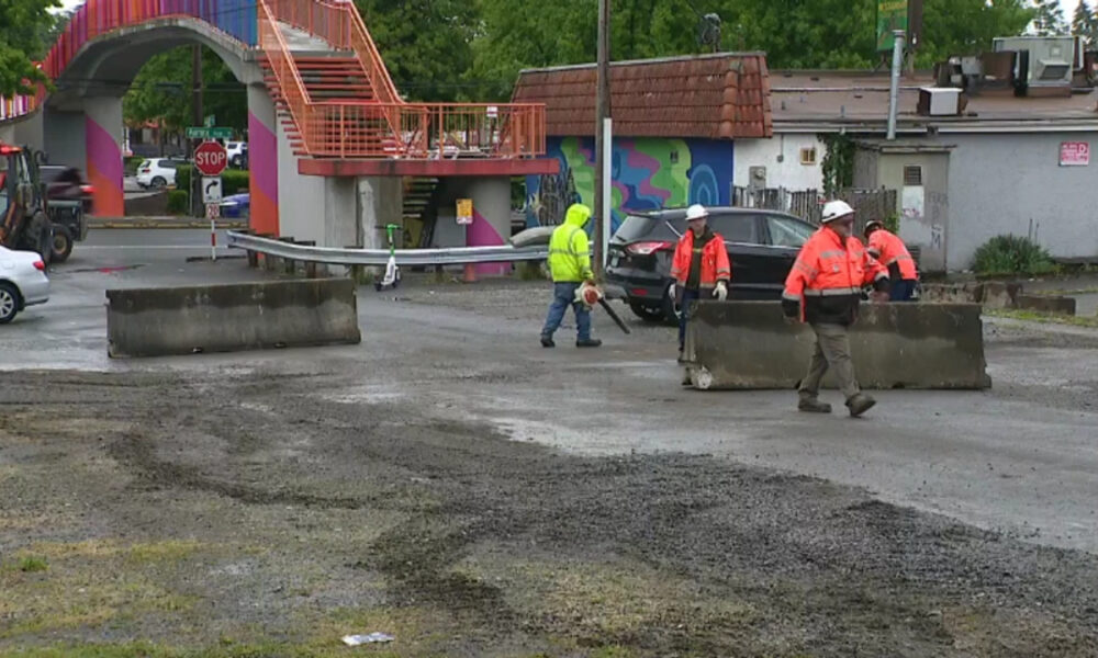 Construction workers in bright orange vests repositioning concrete barriers on a wet street near a colorful curved pedestrian bridge.