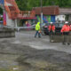 Construction workers in bright orange vests repositioning concrete barriers on a wet street near a colorful curved pedestrian bridge.