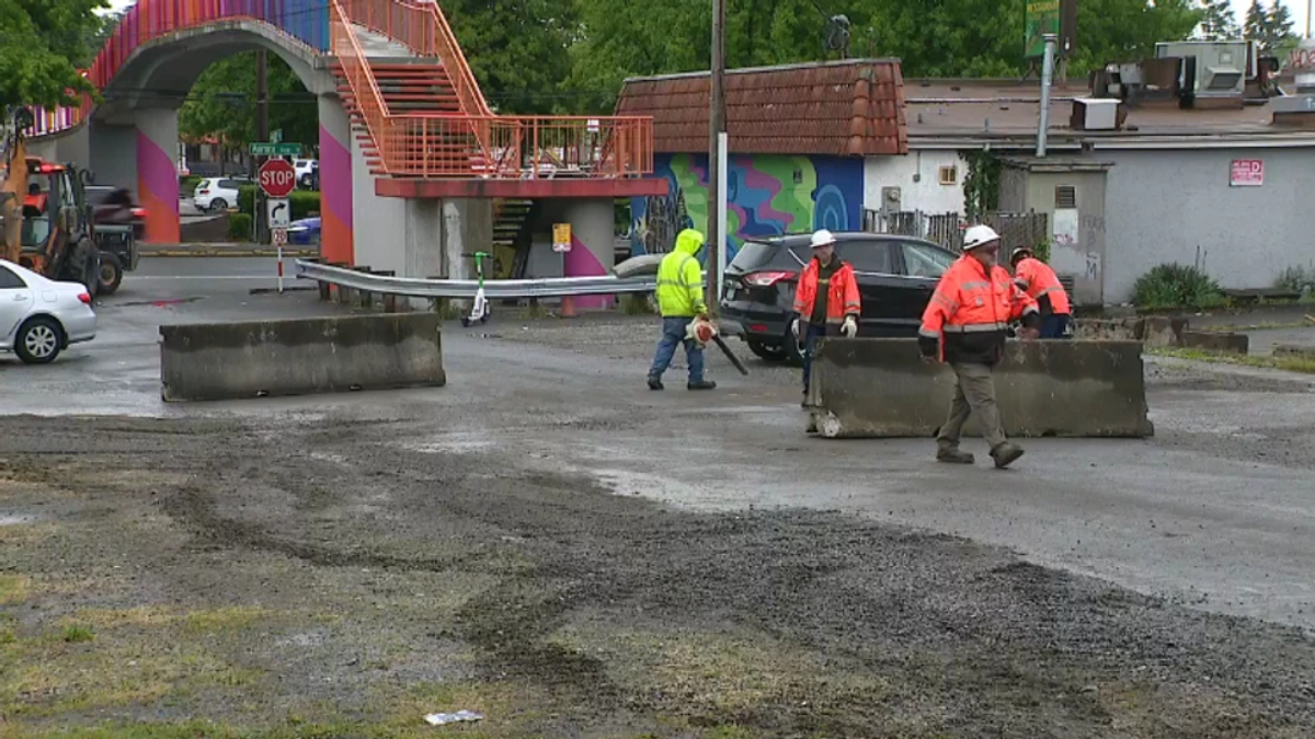Construction workers in bright orange vests repositioning concrete barriers on a wet street near a colorful curved pedestrian bridge.