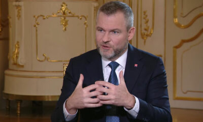 Man in a dark suit and blue tie speaks, hands gesturing in front of an ornate gold-trimmed backdrop.