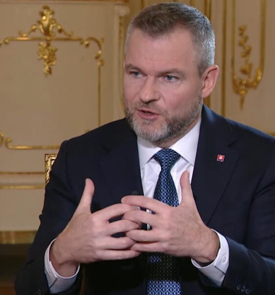 Man in a dark suit and blue tie speaks, hands gesturing in front of an ornate gold-trimmed backdrop.