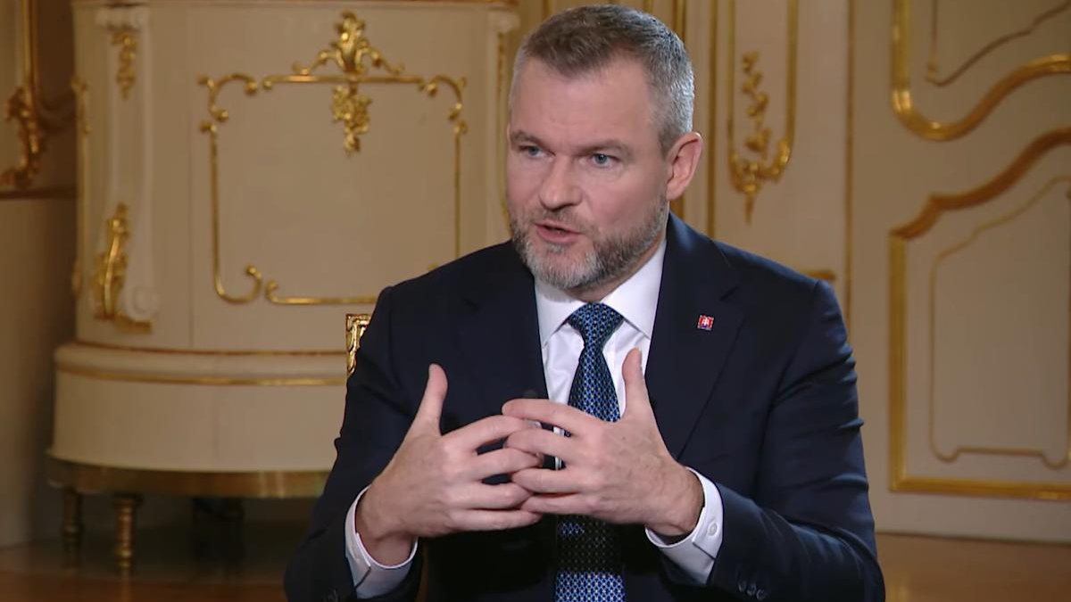 Man in a dark suit and blue tie speaks, hands gesturing in front of an ornate gold-trimmed backdrop.