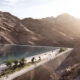 Futuristic elevated walkway over a deep canyon with a calm lake to the left; pedestrians stroll along the path as sunlight shines from the right.