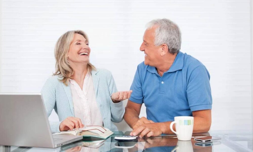 Smiling older couple sits at a glass table with a laptop, notebook, mug, and phone, enjoying a conversation.