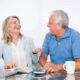 Smiling older couple sits at a glass table with a laptop, notebook, mug, and phone, enjoying a conversation.