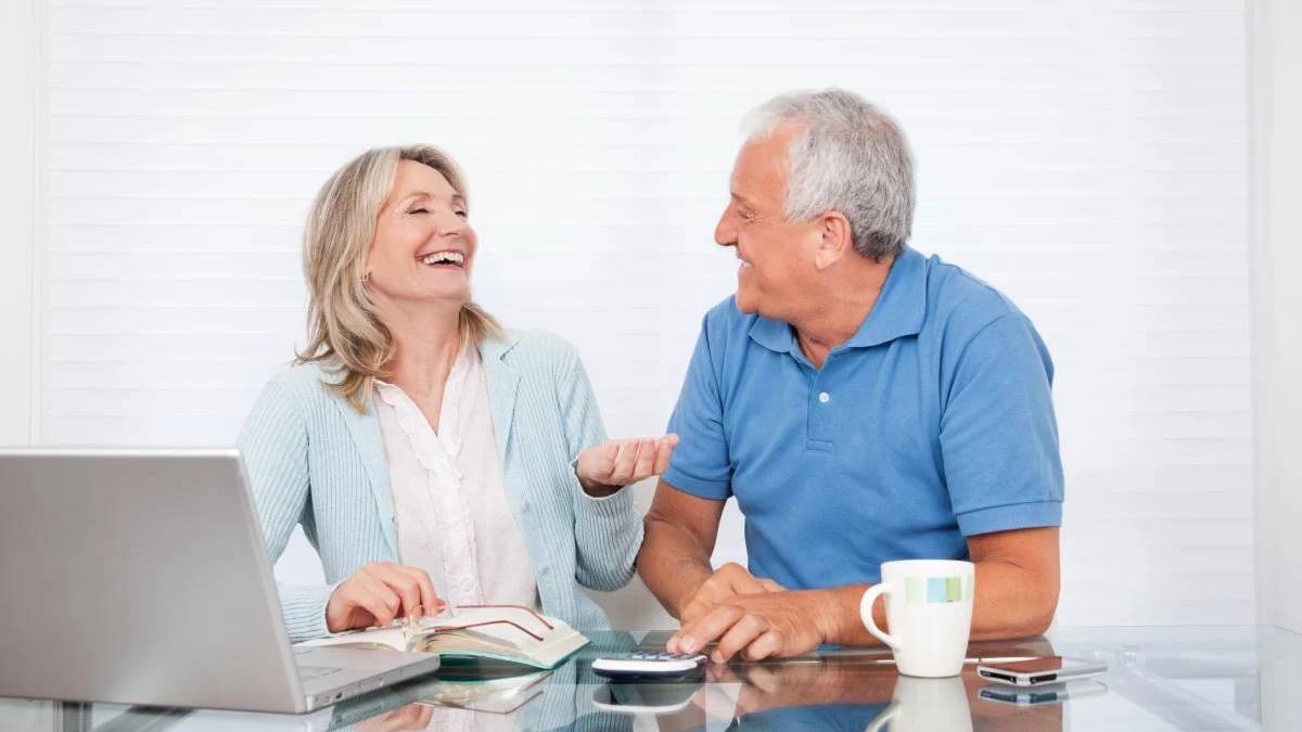 Smiling older couple sits at a glass table with a laptop, notebook, mug, and phone, enjoying a conversation.