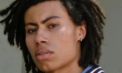 Close-up portrait of a person with dark shoulder-length dreadlocks and a small earring, looking at the camera.