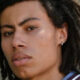 Close-up portrait of a person with dark shoulder-length dreadlocks and a small earring, looking at the camera.
