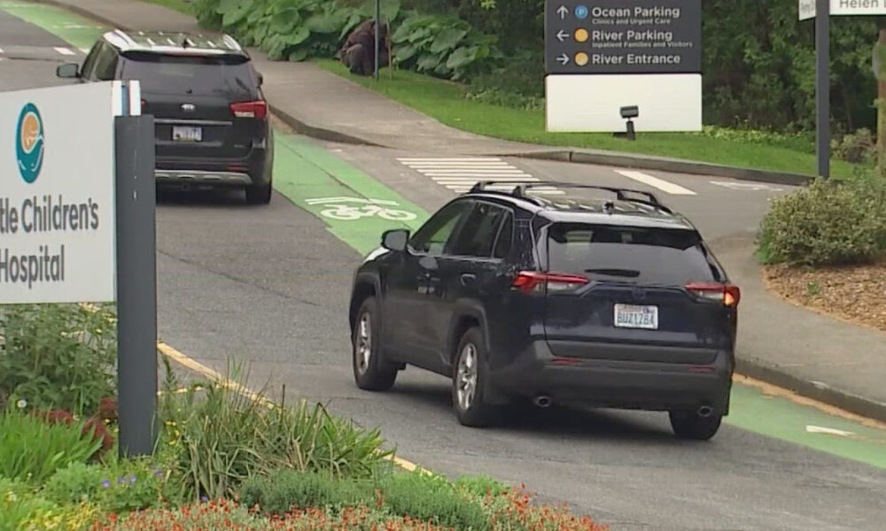 Dark SUV driving in a bike lane near Seattle Children's Hospital, with directional signs in the background.