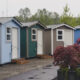 Row of colorful small cabins or sheds with individual doors and step platforms, string lights along the eaves, and potted plants in the foreground