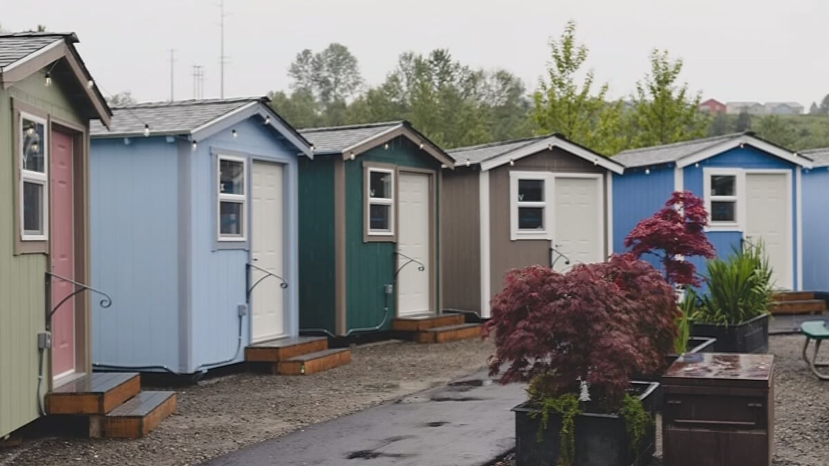 Row of colorful small cabins or sheds with individual doors and step platforms, string lights along the eaves, and potted plants in the foreground