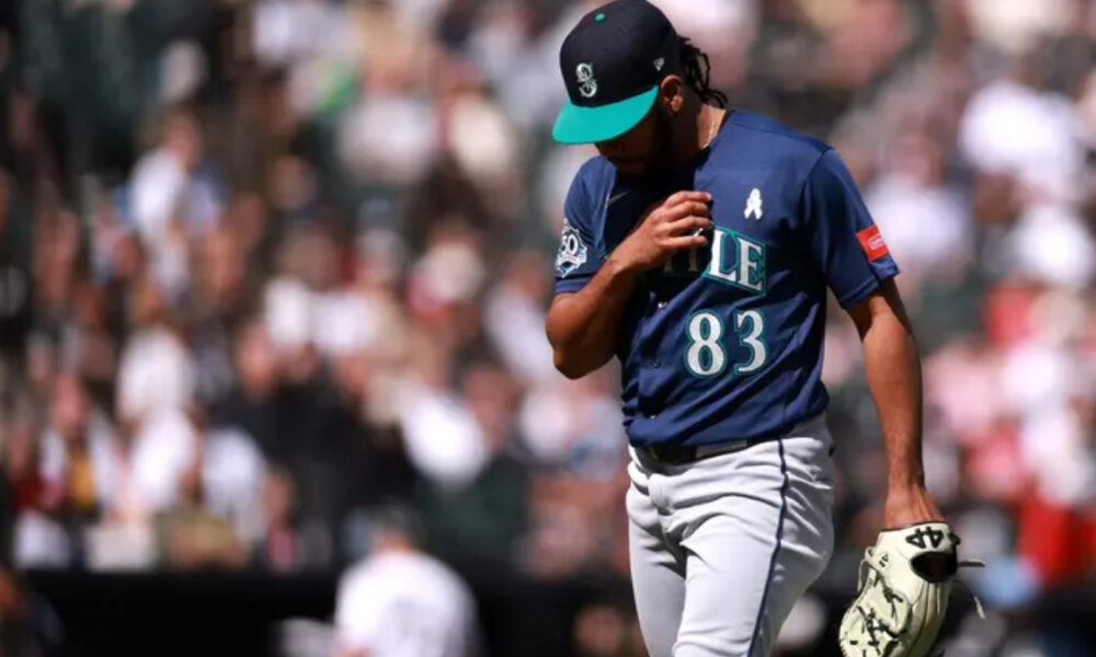 Baseball player in a navy jersey with number 83, teal cap, adjusting his jersey while holding a glove on the field, blurred crowd behind him.