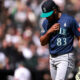 Baseball player in a navy jersey with number 83, teal cap, adjusting his jersey while holding a glove on the field, blurred crowd behind him.