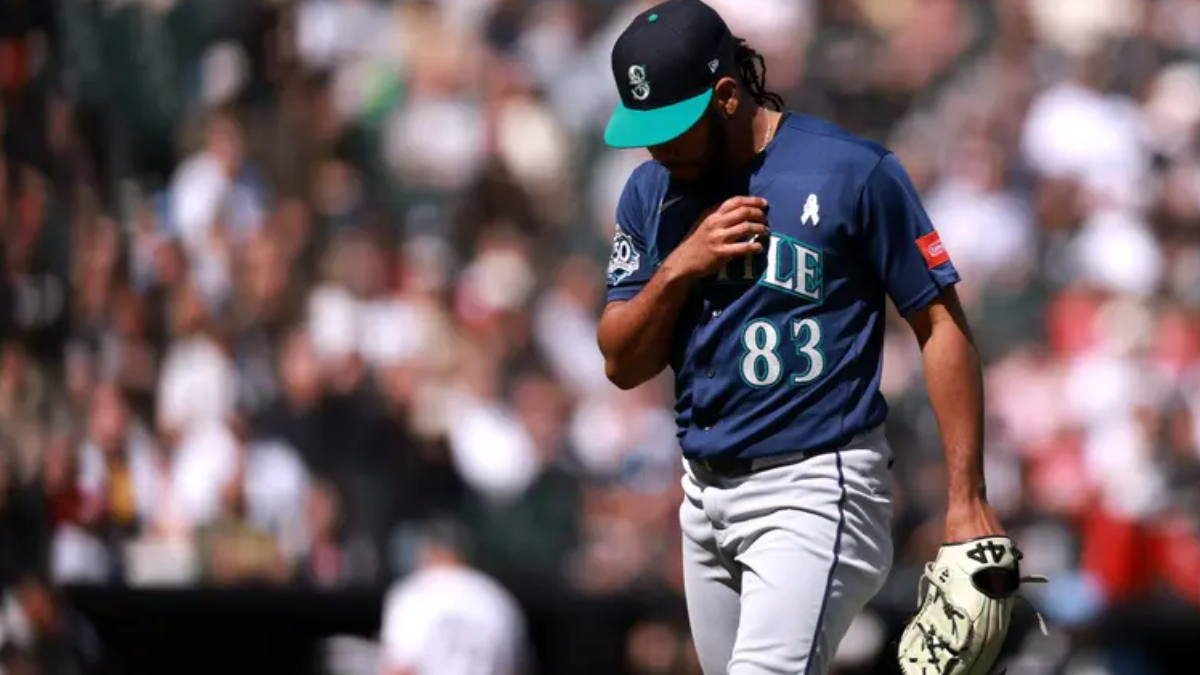 Baseball player in a navy jersey with number 83, teal cap, adjusting his jersey while holding a glove on the field, blurred crowd behind him.