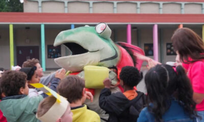 Group of children gathered around a large green and red dinosaur mascot with its mouth open at a school event, colorful building columns in the background.