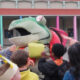 Group of children gathered around a large green and red dinosaur mascot with its mouth open at a school event, colorful building columns in the background.