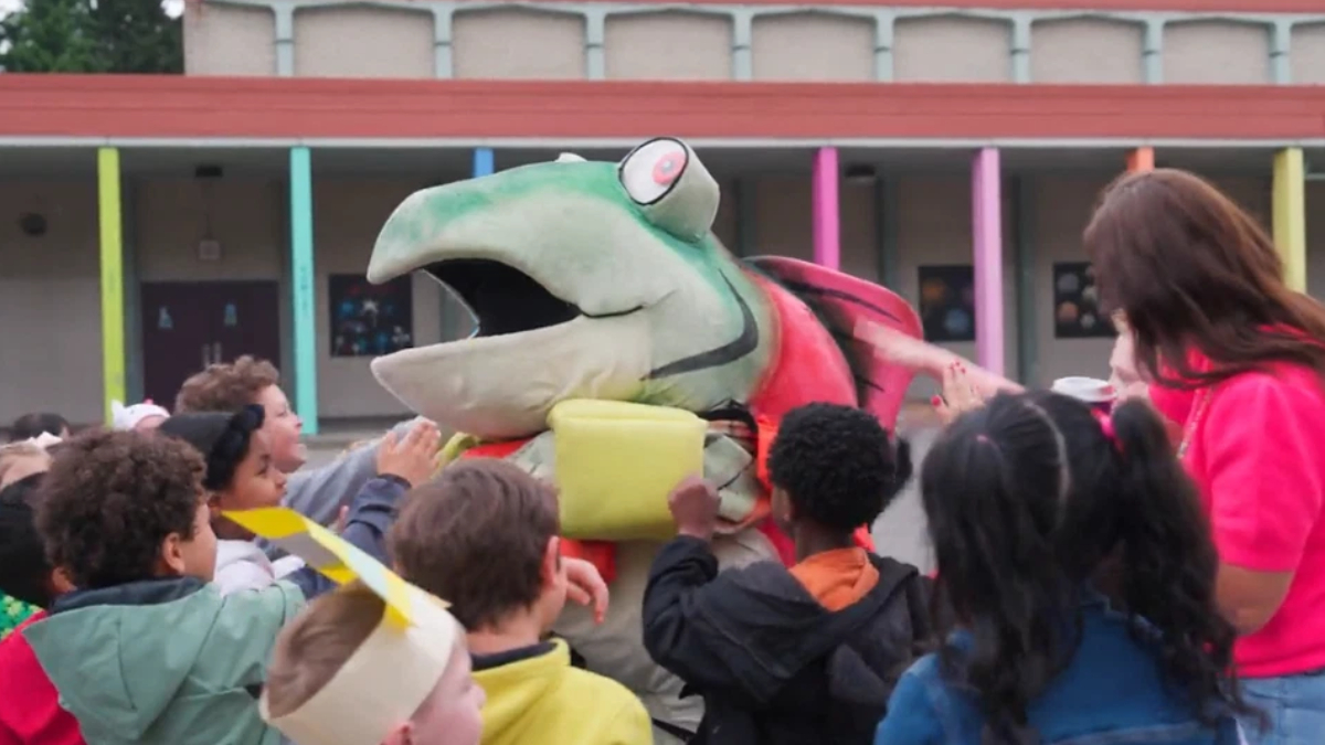 Group of children gathered around a large green and red dinosaur mascot with its mouth open at a school event, colorful building columns in the background.