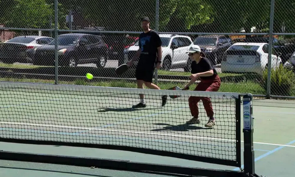 Two people play pickleball on an outdoor court as a neon ball arcs toward the net under a chain-link fence.