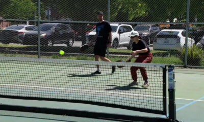 Two people play pickleball on an outdoor court as a neon ball arcs toward the net under a chain-link fence.