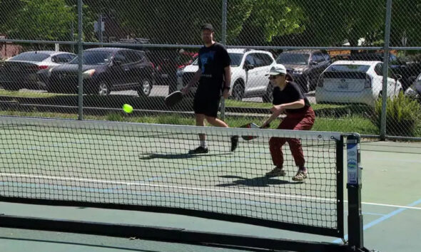 Two people play pickleball on an outdoor court as a neon ball arcs toward the net under a chain-link fence.
