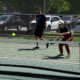 Two people play pickleball on an outdoor court as a neon ball arcs toward the net under a chain-link fence.