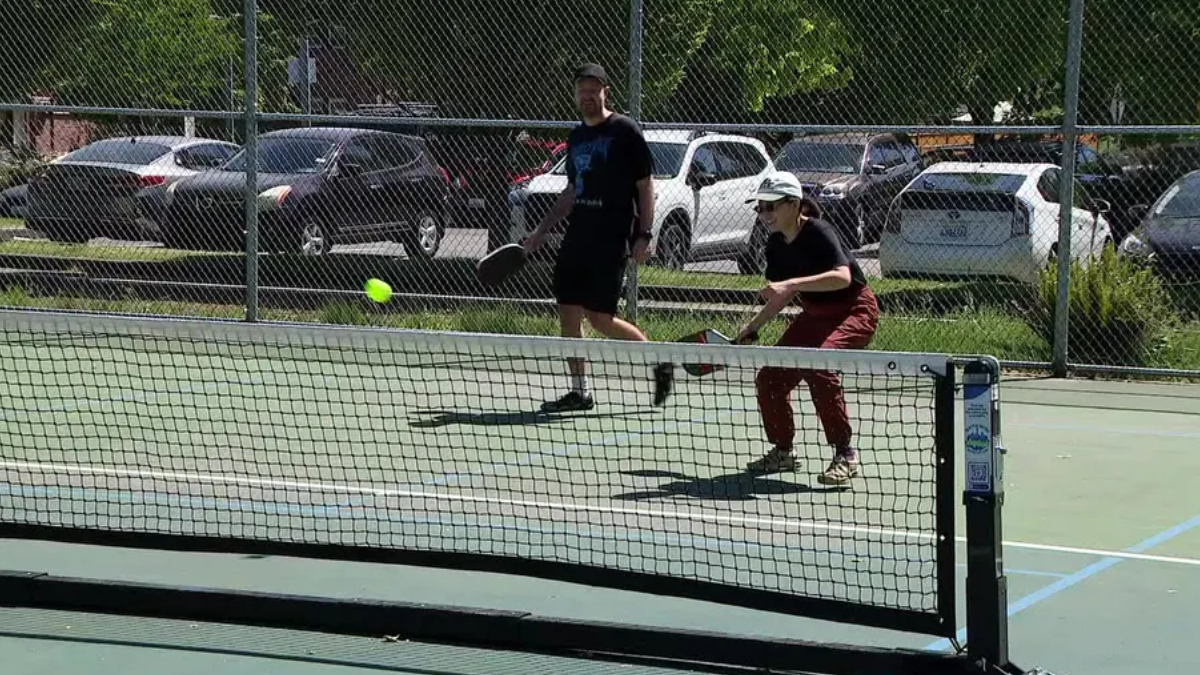 Two people play pickleball on an outdoor court as a neon ball arcs toward the net under a chain-link fence.