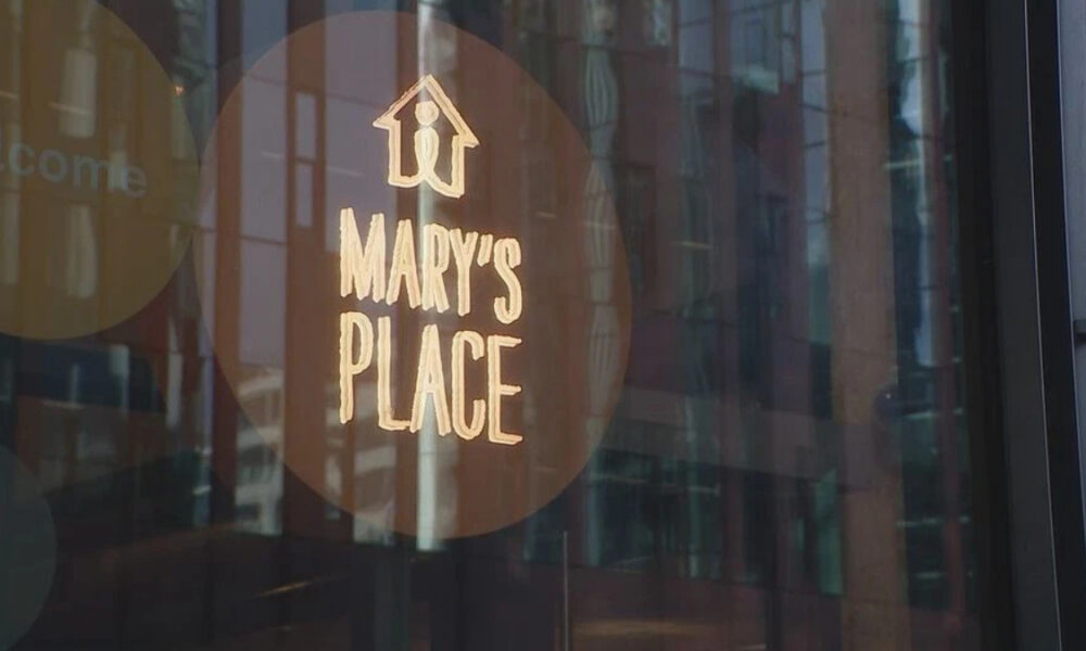 Circular brown sticker on a glass storefront reading 'MARY'S PLACE' with a small house icon above; city reflections visible in the glass.