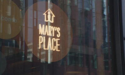 Circular brown sticker on a glass storefront reading 'MARY'S PLACE' with a small house icon above; city reflections visible in the glass.