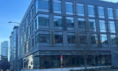 Corner view of a modern glass-and-metal office building along a city street, with a leafless tree in front and blue sky above.