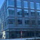 Corner view of a modern glass-and-metal office building along a city street, with a leafless tree in front and blue sky above.