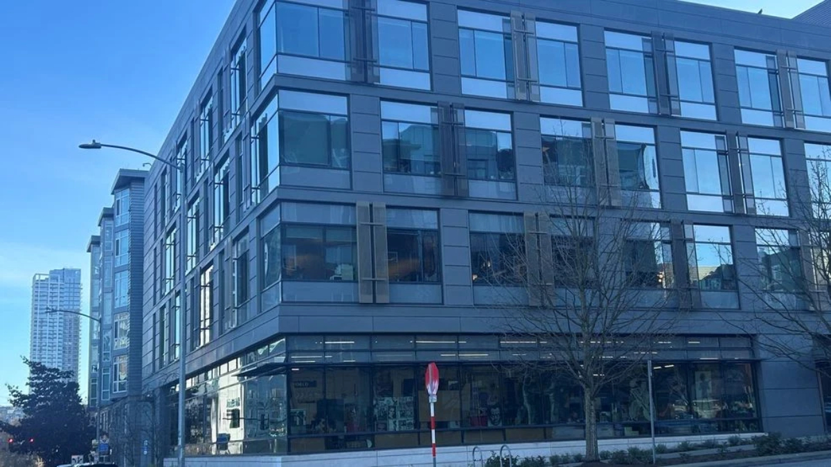 Corner view of a modern glass-and-metal office building along a city street, with a leafless tree in front and blue sky above.