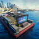 Floating water-stage with a large crowd watching a soccer celebration event by the city skyline and harbor.