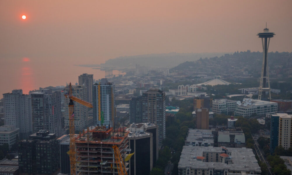City skyline at sunset with an orange-hued sky, a construction crane in the foreground, and the Space Needle to the right over a coastal city.