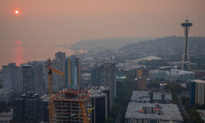 City skyline at sunset with an orange-hued sky, a construction crane in the foreground, and the Space Needle to the right over a coastal city.