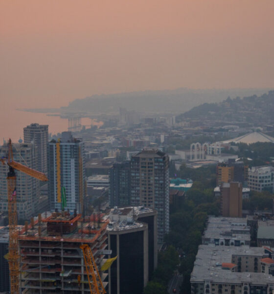 City skyline at sunset with an orange-hued sky, a construction crane in the foreground, and the Space Needle to the right over a coastal city.
