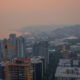 City skyline at sunset with an orange-hued sky, a construction crane in the foreground, and the Space Needle to the right over a coastal city.