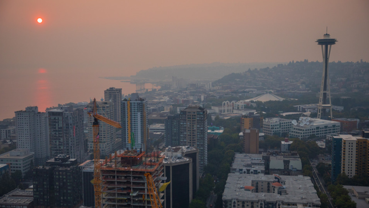 City skyline at sunset with an orange-hued sky, a construction crane in the foreground, and the Space Needle to the right over a coastal city.