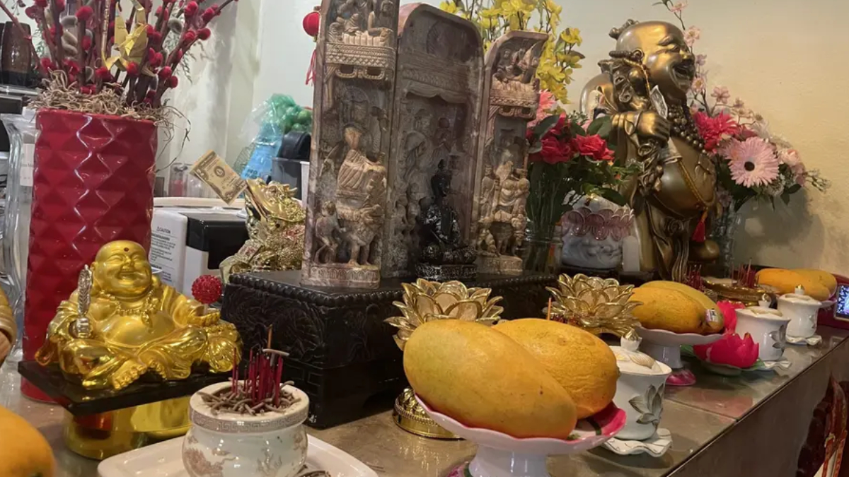 Altar setup with a golden laughing Buddha, incense, flowers, and large mango offerings on the table.