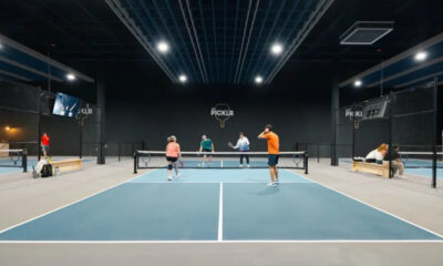 Indoor pickleball court with four players near the net; a player in orange serves as others watch, PICKLR logo on the back wall.